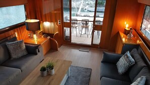 Un intérieur chaleureux avec un canapé, une table en bois agrémentée de plantes, et une vue sur la terrasse et l’eau à l’extérieur.