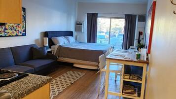 A compact hotel room with a kitchenette, a sofa, a bed, a dining area, and a view through the window.