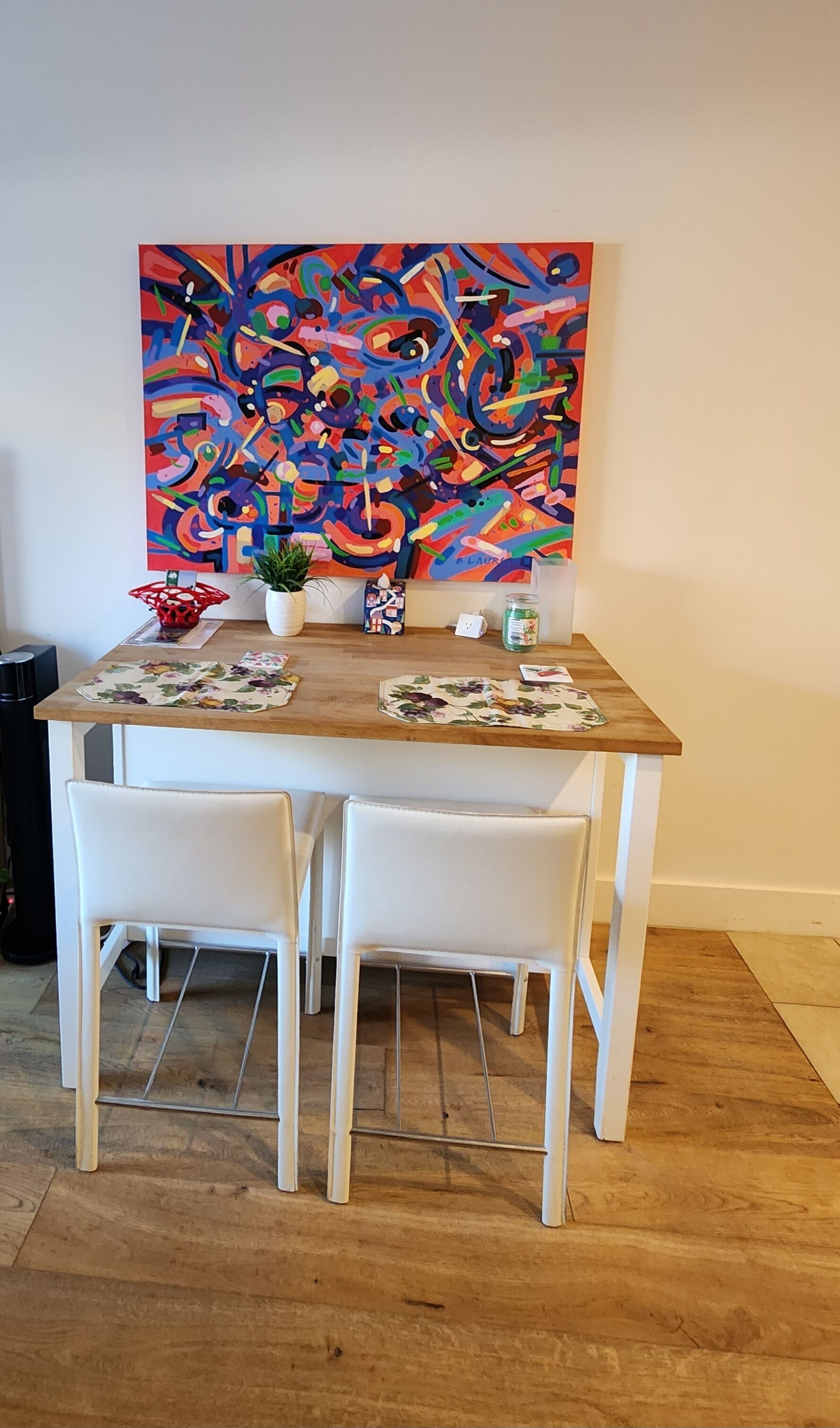 A dining area with a wooden table, two white chairs, a colorful abstract painting on the wall, and a small potted plant.