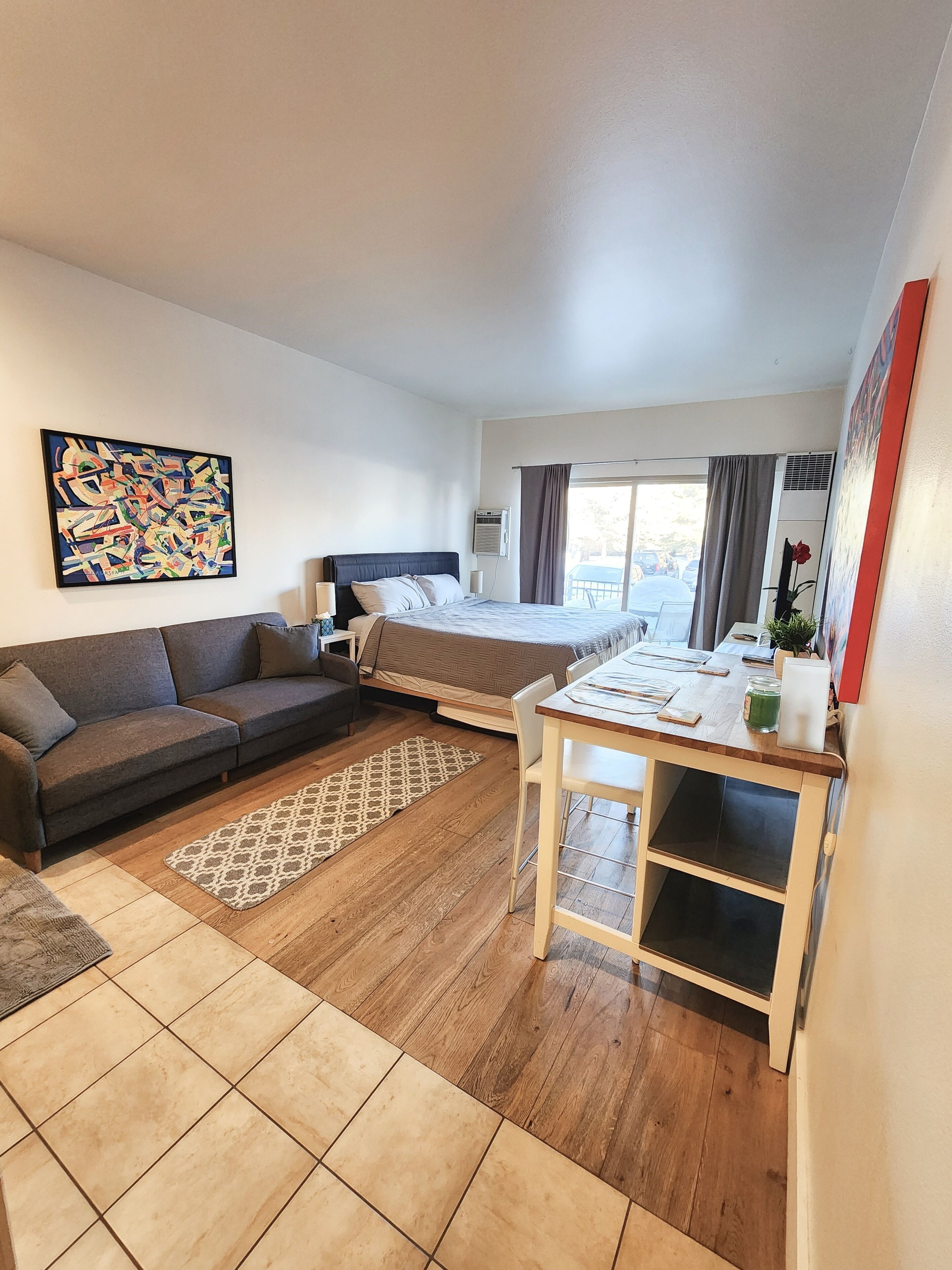 A modern hotel room with a bed, a sofa, a dining table, and a large window.