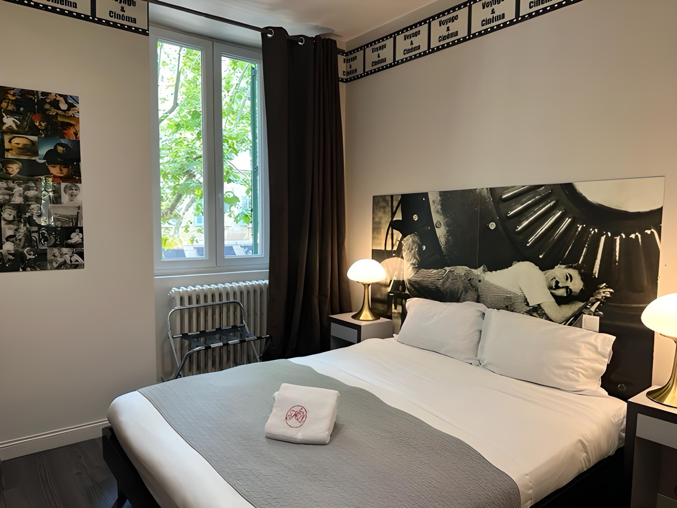A hotel room with a large bed, two bedside lamps, a radiator, a window with curtains, and a wall with a film strip design.
