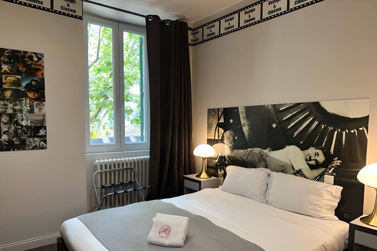 Quarto de hotel com uma cama grande, duas luminárias de cabeceira, um radiador, uma janela com cortinas e uma parede com design de faixa de filme.