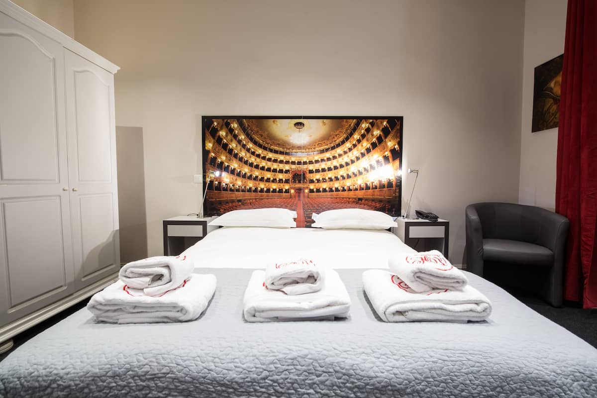 Quarto de hotel com uma cama grande, um quadro na parede, uma cadeira e uma escrivaninha com abajur.