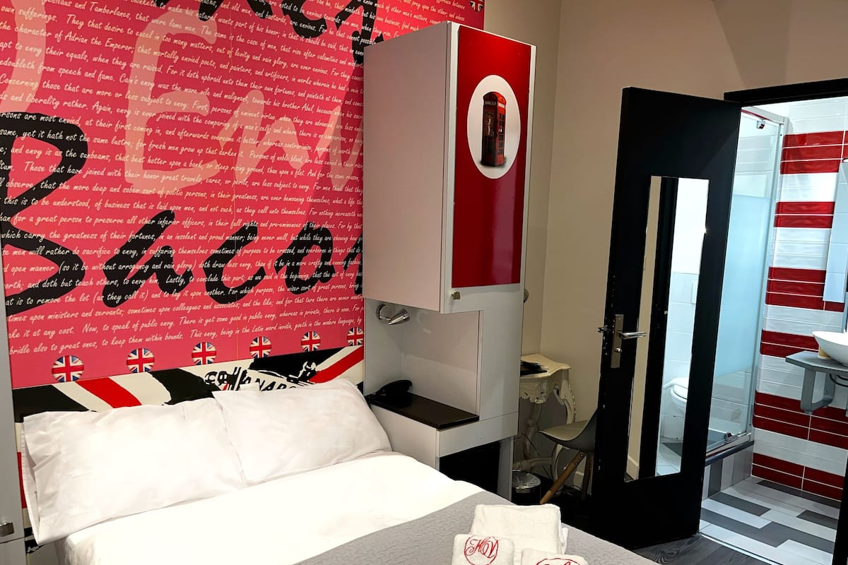Quarto de hotel com uma cama, parede vermelha e branca, uma porta e um banheiro visível através de uma porta aberta.