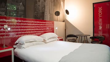Hotelkamer met een groot bed, een bureau met lamp, een stoel, een klein tafeltje en een rode wand met een stadsgezicht muurdecoratie.
