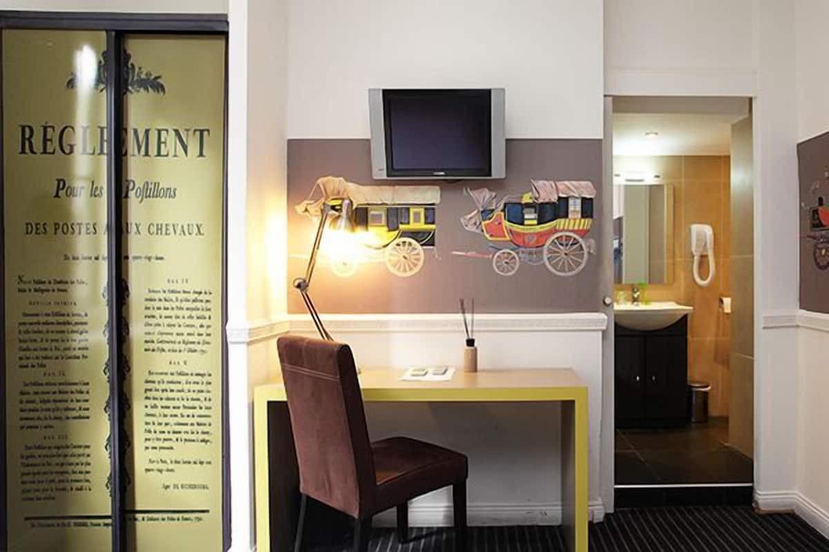 Quarto de hotel com escrivaninha, cadeira e televisão fixada na parede.