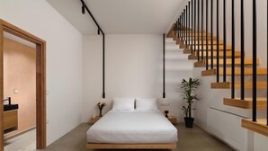 A modern bedroom with a bed centered in the room, a wooden staircase to the right, and a small bedside table with a plant. - Iviskos - Island View (Ιαλυσός)