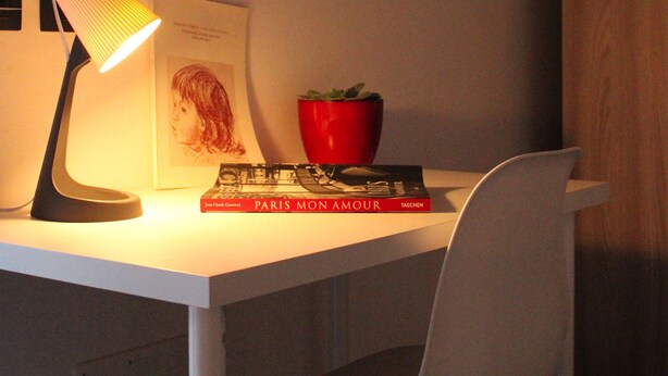 Un bureau avec une lampe, un livre intitulé « PARIS, MON AMOUR », un pot rouge et un tableau encadré accroché au mur.