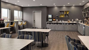 Un espace repas moderne avec des chaises et des tables à rayures, une cuisine dotée de meubles gris et une vue sur l’extérieur grâce à de grandes fenêtres.