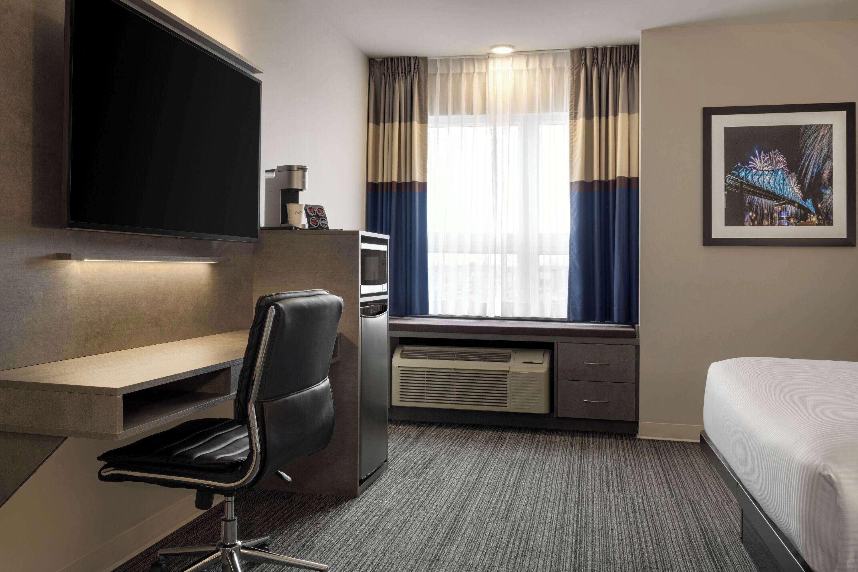 A hotel room with a flat-screen TV mounted on the wall, a desk with a chair, a bed, a window with curtains, and a framed picture on the wall.