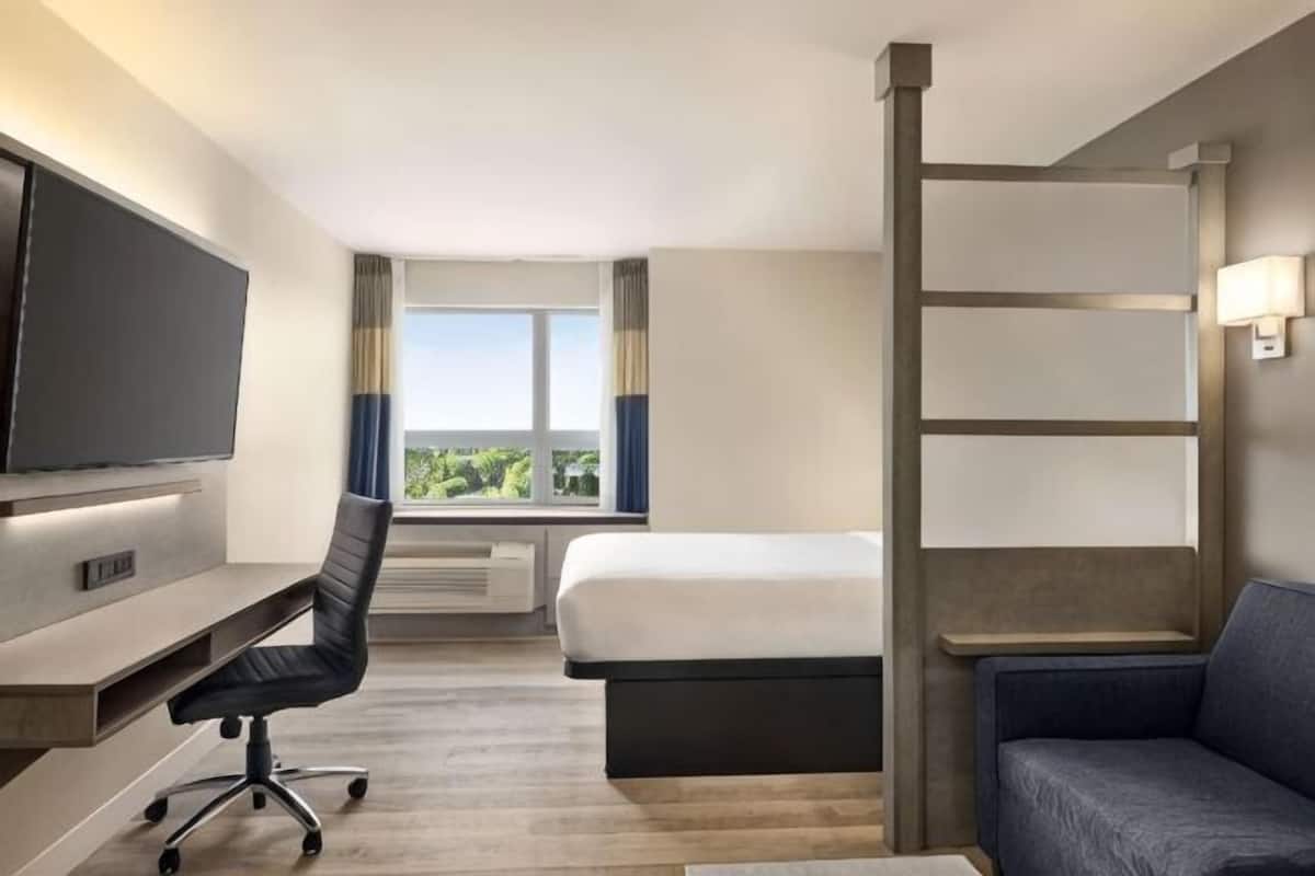 Une chambre d’hôtel comprenant un lit, un bureau avec une chaise, une télévision, une fenêtre avec des rideaux et une vue sur la verdure.
