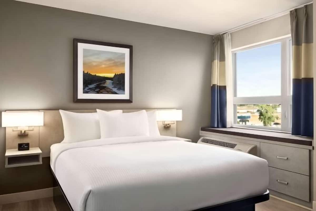Une chambre d’hôtel avec un grand lit, deux tables de chevet avec des lampes, une fenêtre avec des rideaux et un tableau encadré au mur.