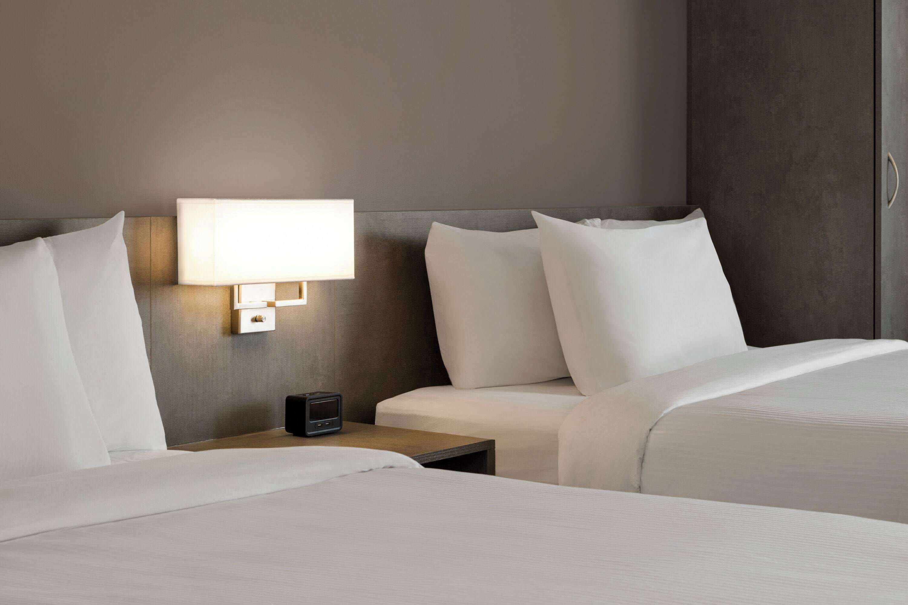 A hotel room with two beds, a bedside table with a lamp, and a small electronic device on the table.