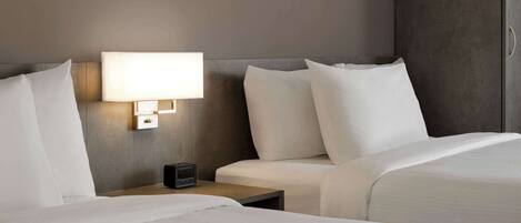 Habitación de hotel con dos camas, una mesita de noche con lámpara y un pequeño dispositivo electrónico sobre la mesa.
