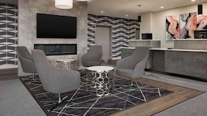 Un hall d’hôtel moderne avec un grand téléviseur, des fauteuils gris, une table basse ronde et un tapis à motifs géométriques.