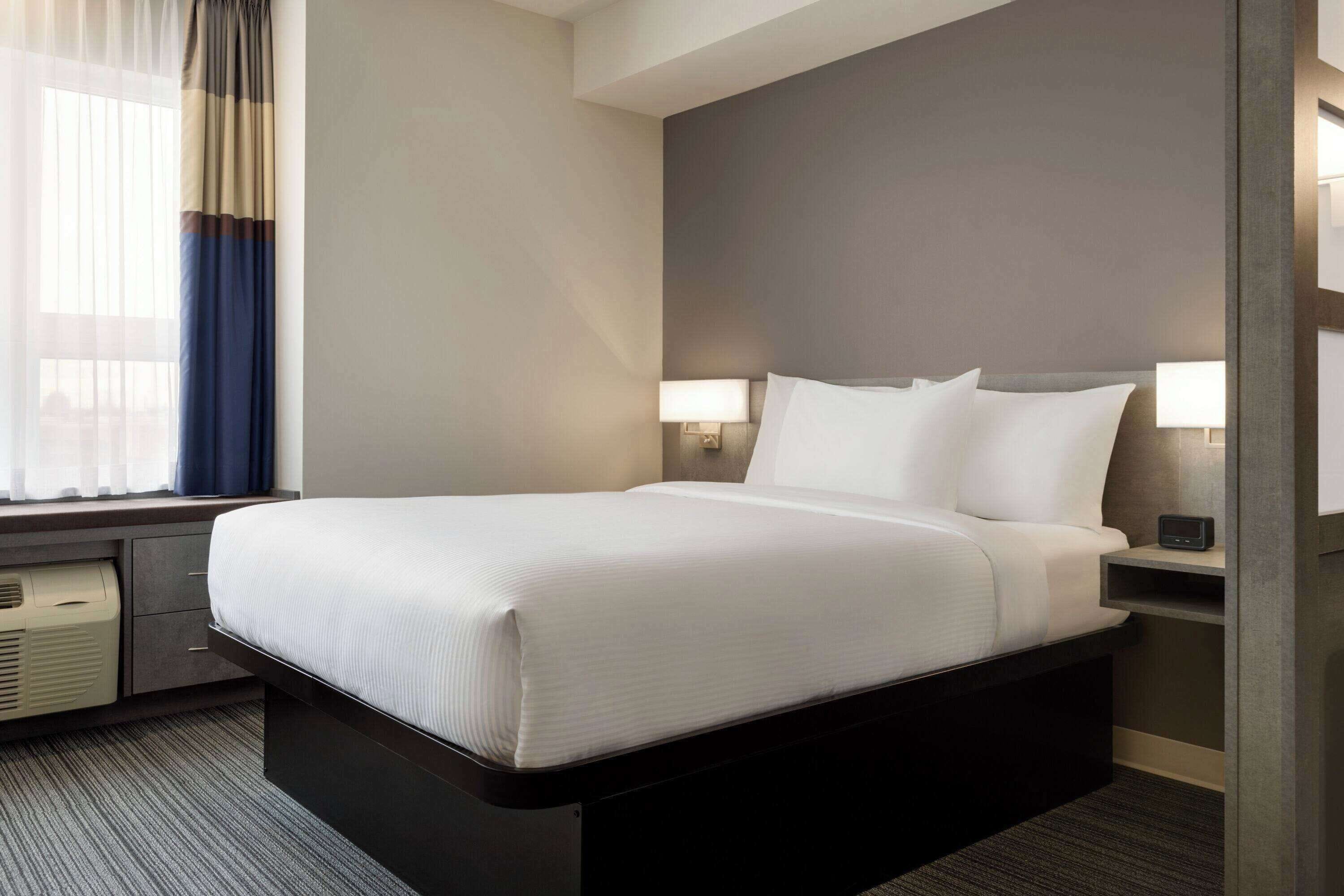 A hotel room with a large bed, bedside lamps, a window with curtains, and a small heater.