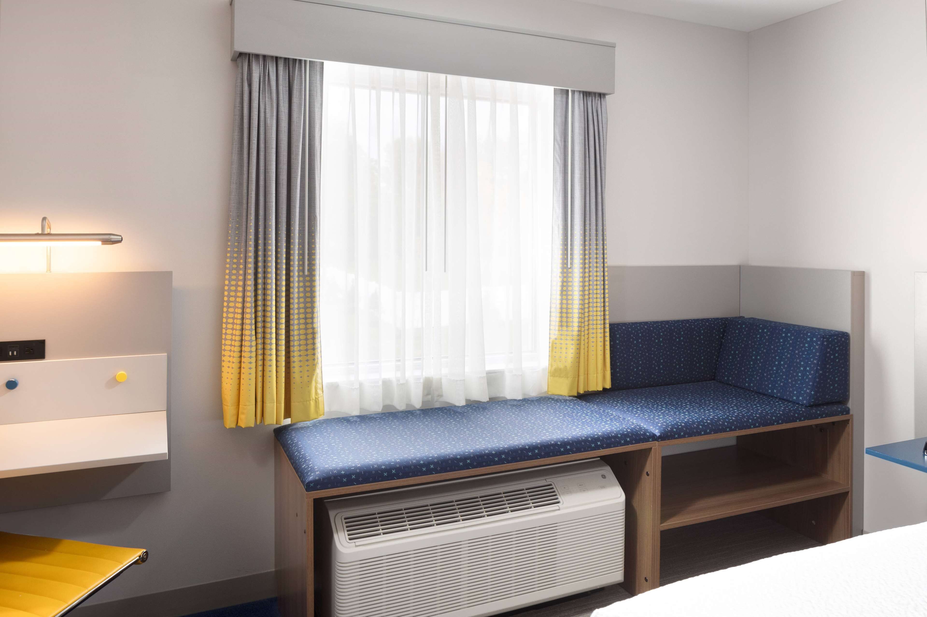 A hotel room with a corner bed, a bedside table, a lamp, a window with curtains, and a built-in heater.