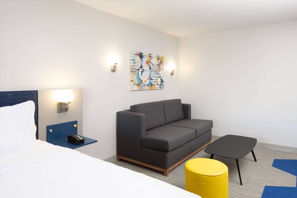 A modern hotel room with a bed, a gray sofa, a small table, and a yellow ottoman.