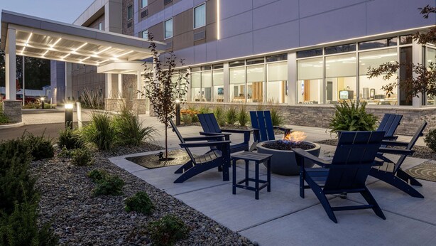 An outdoor seating area with Adirondack chairs and a fire pit, surrounded by a landscaped garden and a building with large windows.