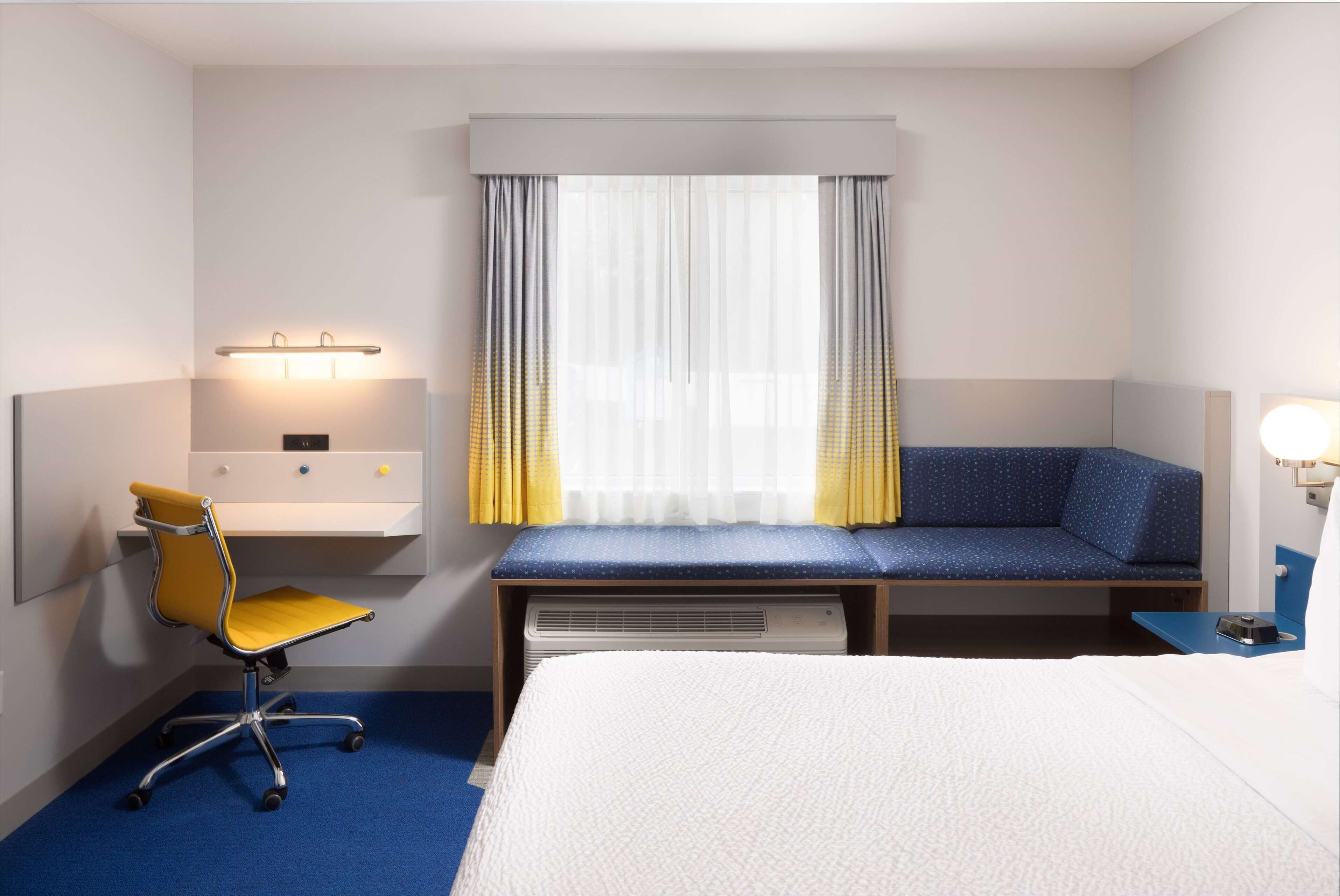 Una habitación de hotel con una cama, un escritorio con silla, una ventana con cortinas y una mesita de noche con lámpara.