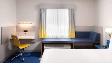Una habitación de hotel con una cama, un escritorio con silla, una ventana con cortinas y una mesita de noche con lámpara.