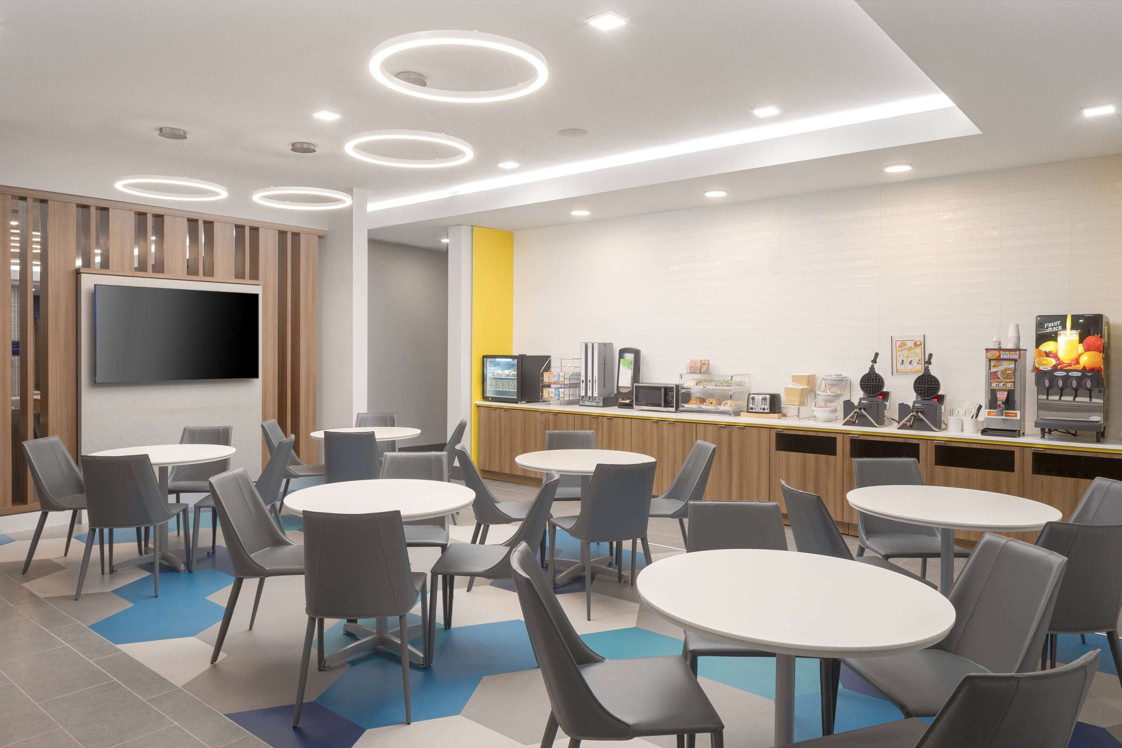 Un espace repas moderne avec des tables et des chaises rondes, un téléviseur à écran plat et un comptoir équipé d’une machine à café et d’un présentoir pour les aliments.