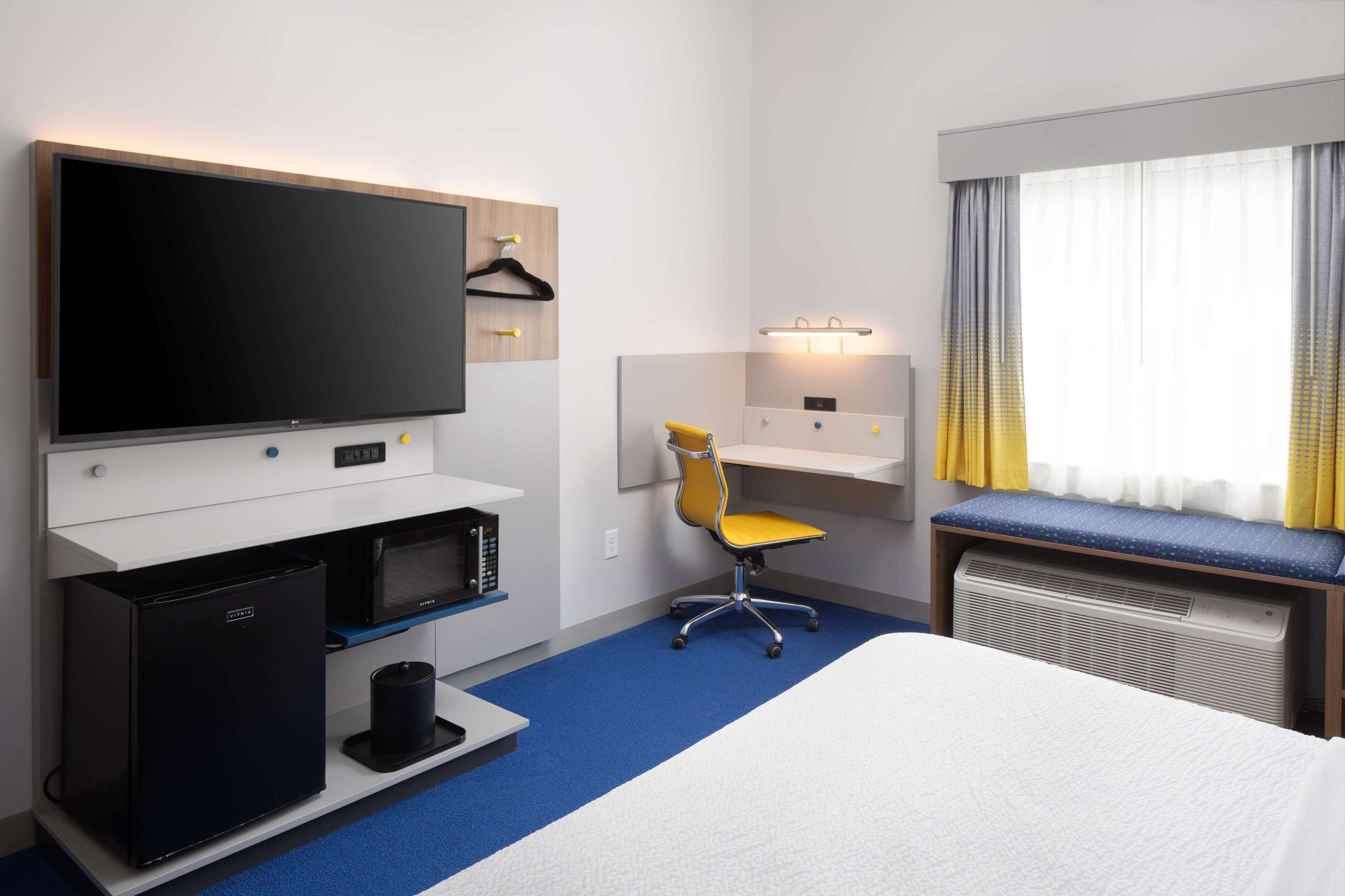 Hotelkamer met een groot flatscreen televisietoestel, een bureau met stoel, een bed, een kleine kitchenette en een raam met gordijnen.