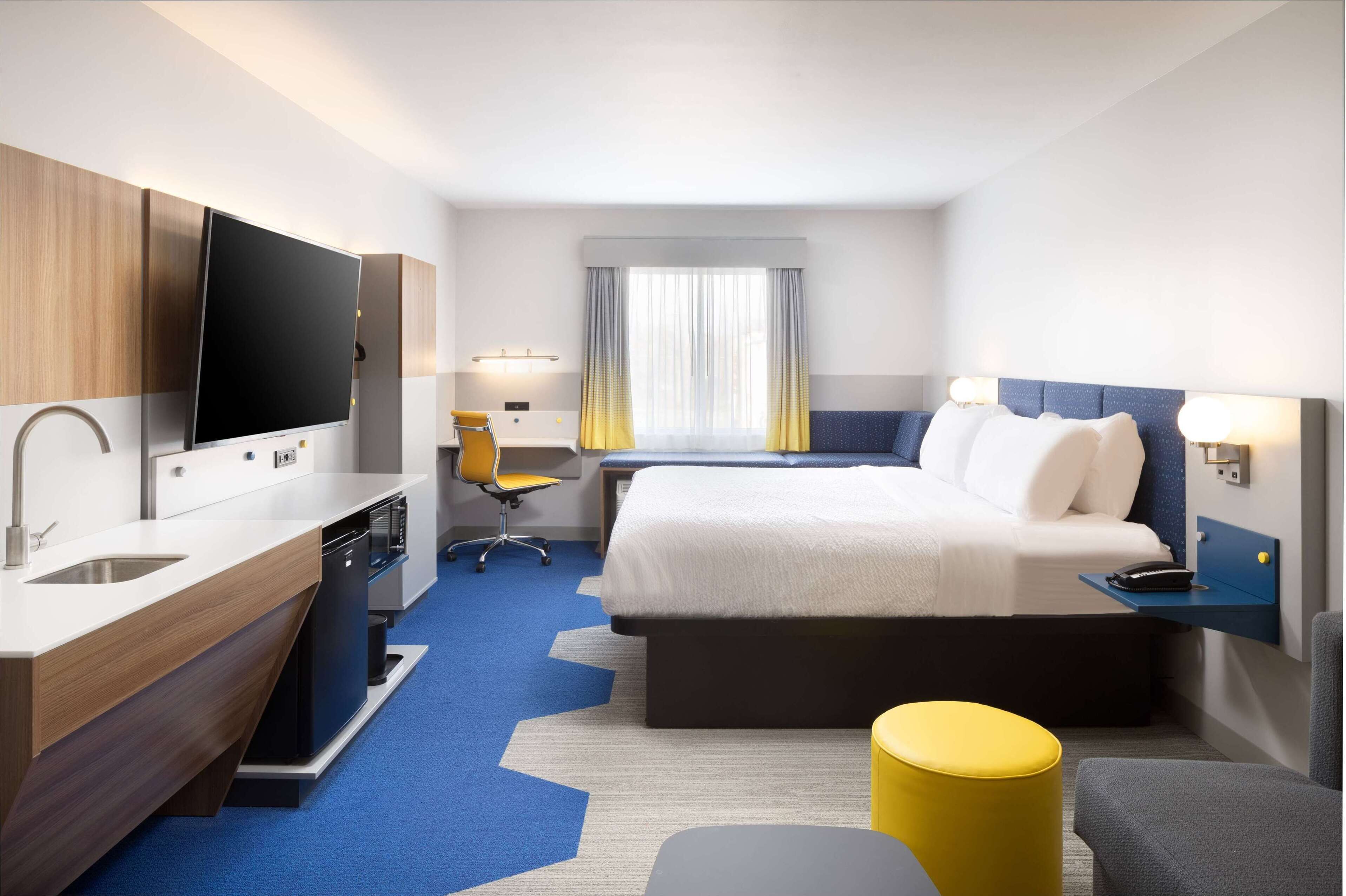 Una habitación de hotel con una cama, un escritorio con silla, un televisor, un lavamanos y una ventana con cortinas.