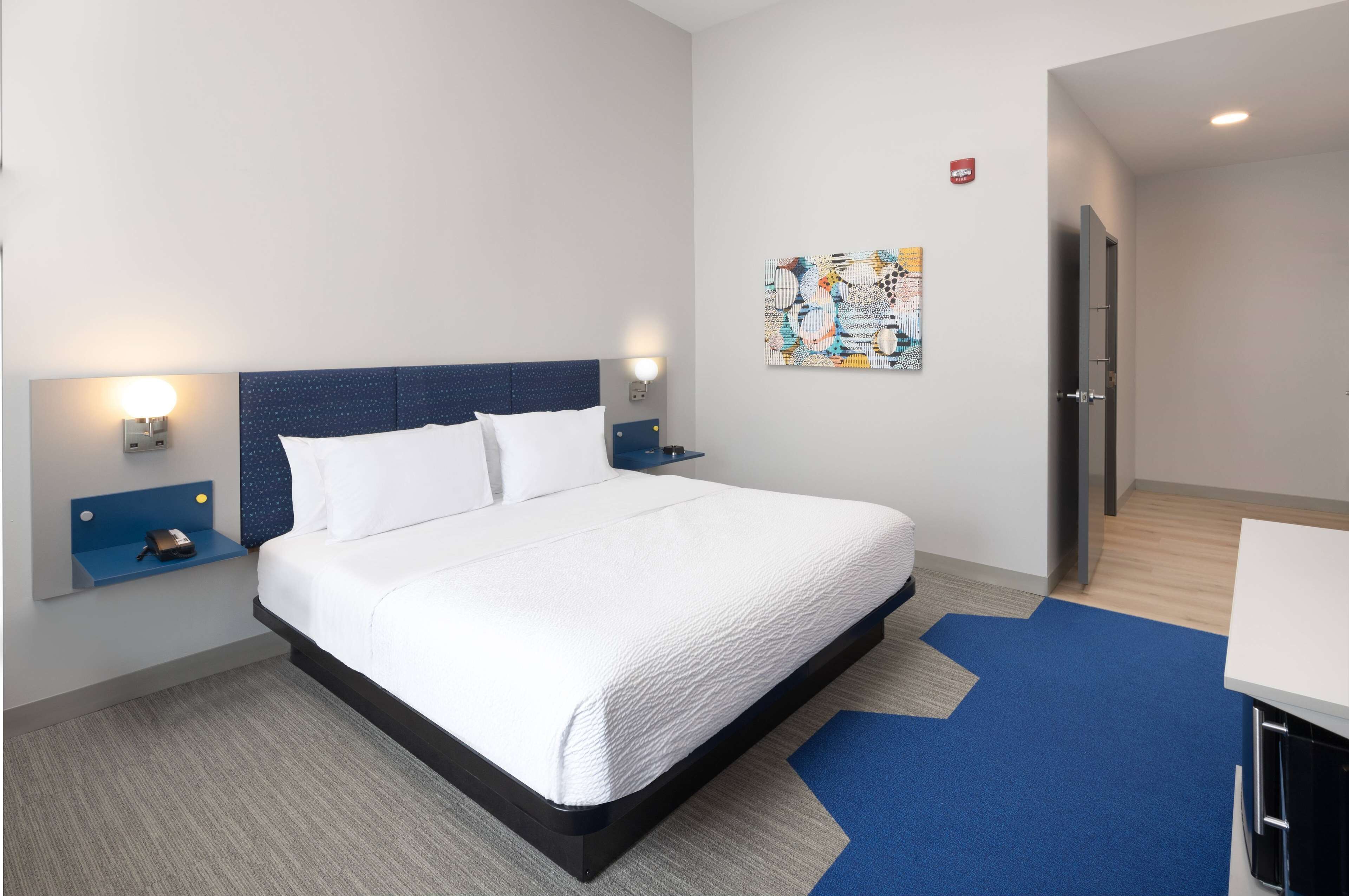 A modern hotel room with a large bed, a nightstand, a wall-mounted lamp, and a painting.