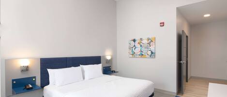 A modern hotel room with a large bed, a nightstand, a wall-mounted lamp, and a painting.