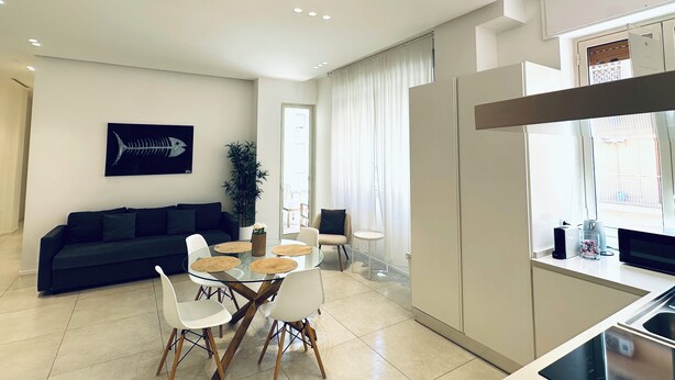 A modern living room with a dark sofa, a glass dining table with white chairs, and a kitchen area with a refrigerator and microwave. - Fantastic Apartment in Historic Center - Palermo (Palermo)