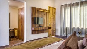 A modern hotel room with a large bed, a flat-screen TV mounted on a wooden panel, a desk with a chair, and a view of the outdoors through sheer curtains. - MAULIK RETREAT (Ramnagar)