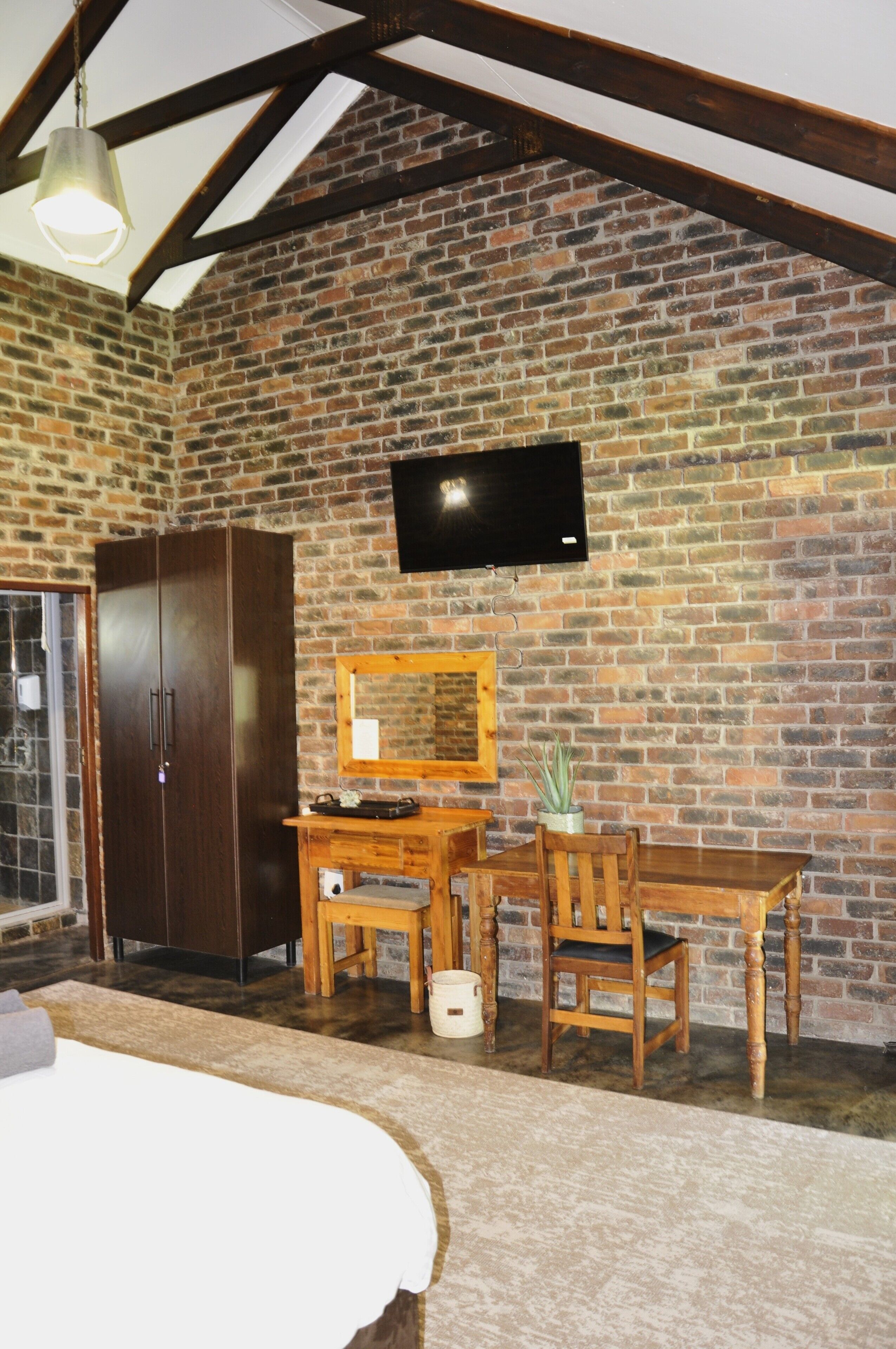 A room with a brick wall, a wooden wardrobe, a television mounted on the wall, a wooden dining table with chairs, and a bed with a white bedspread.