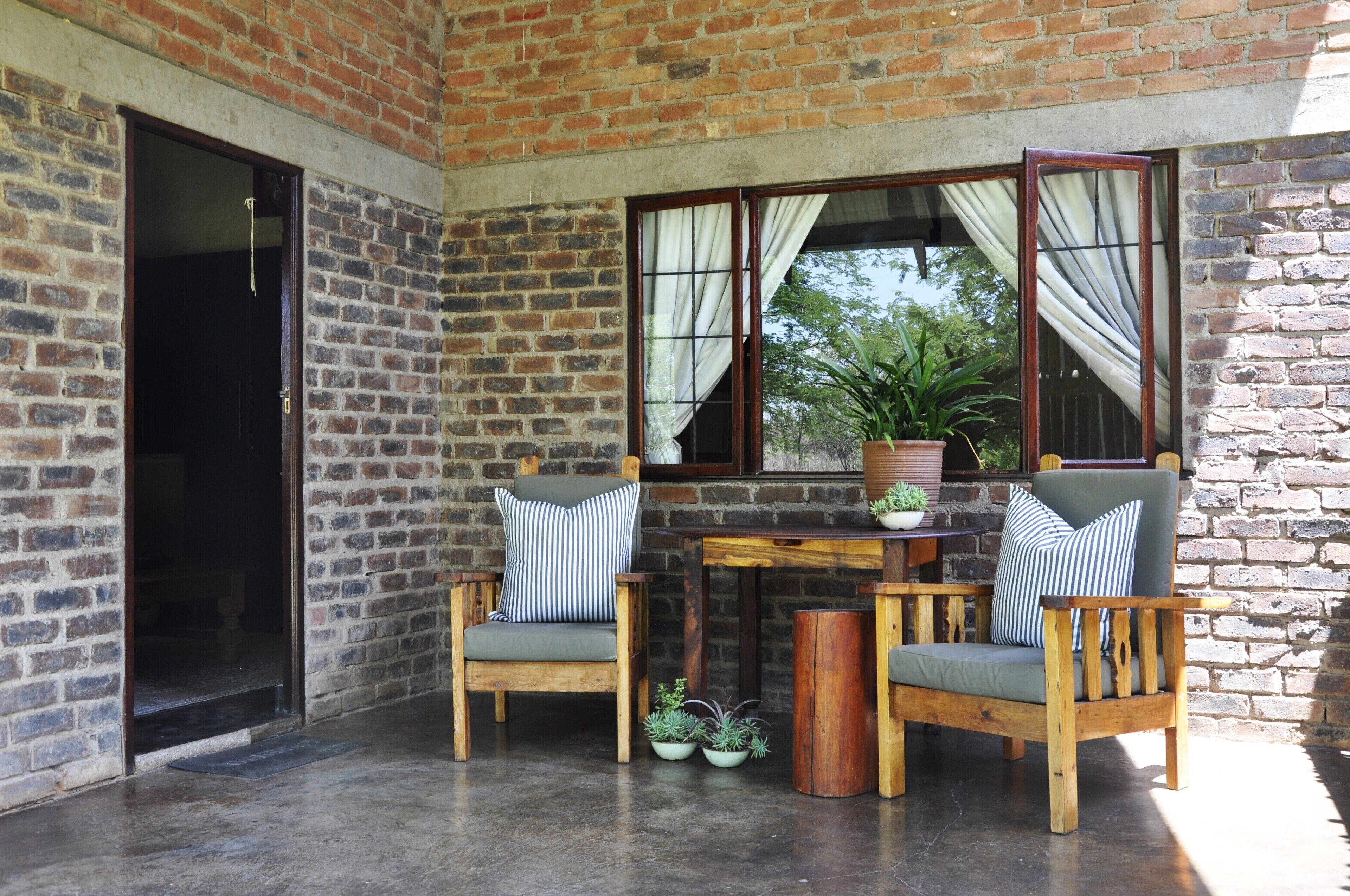A brick-walled room with a wooden table and two chairs, a potted plant, and a window with curtains.