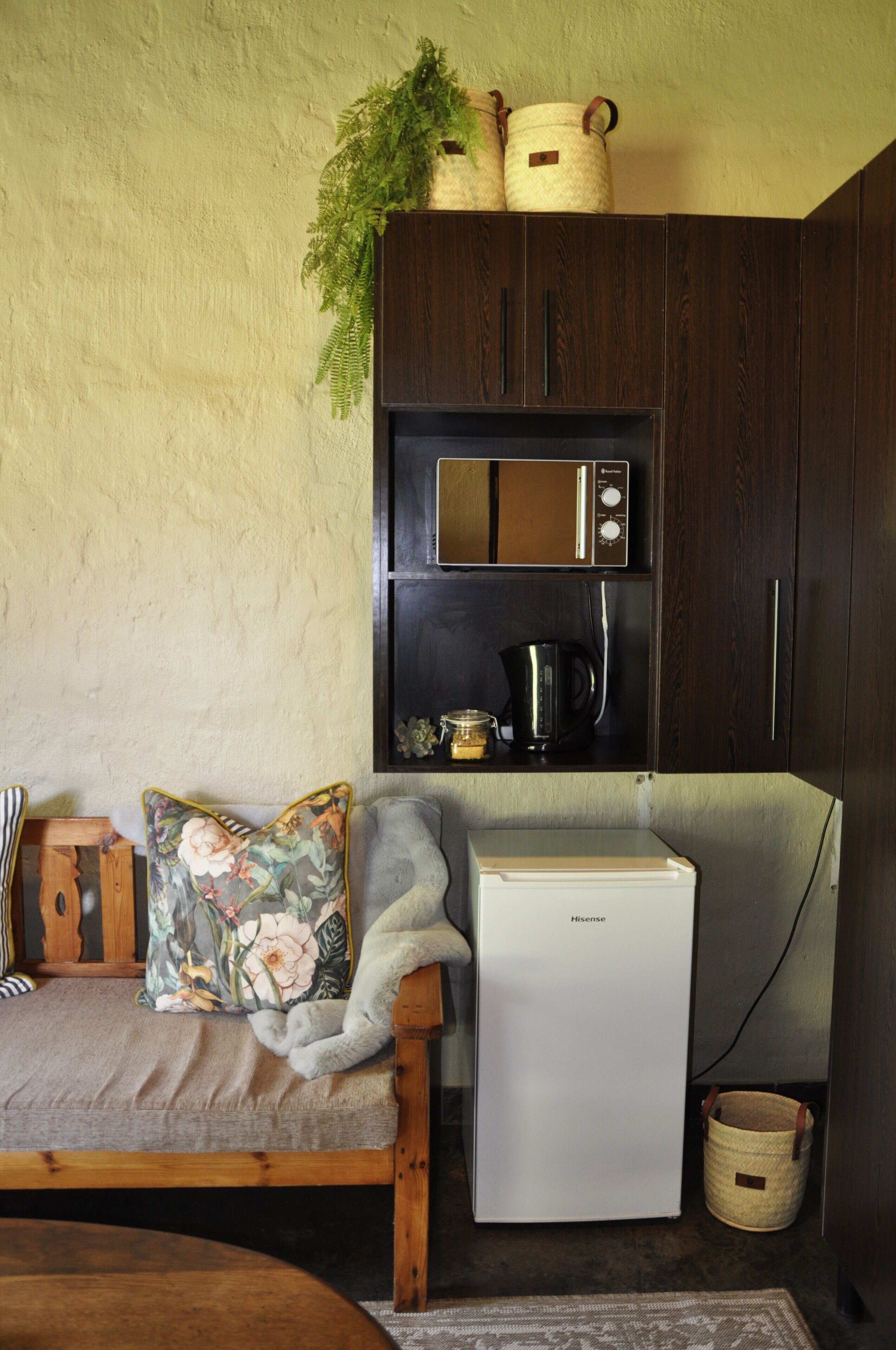 A compact kitchen with a microwave, a small refrigerator, and a sink. There is a wooden bench with a floral cushion and a plant on top of the cabinets.