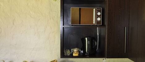 A compact kitchen with a microwave, a small refrigerator, and a sink. There is a wooden bench with a floral cushion and a plant on top of the cabinets.