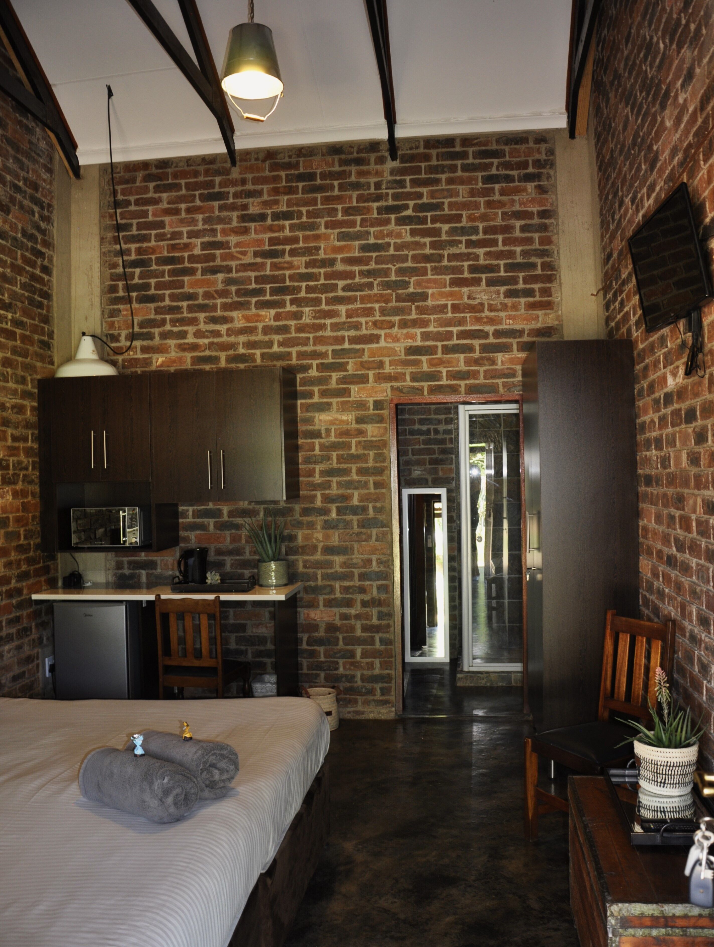 A room with a brick wall, a bed with rolled towels, a small kitchenette, and a door leading to another room.