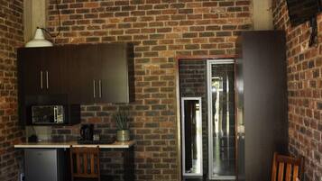 A room with a brick wall, a bed with rolled towels, a small kitchenette, and a door leading to another room.