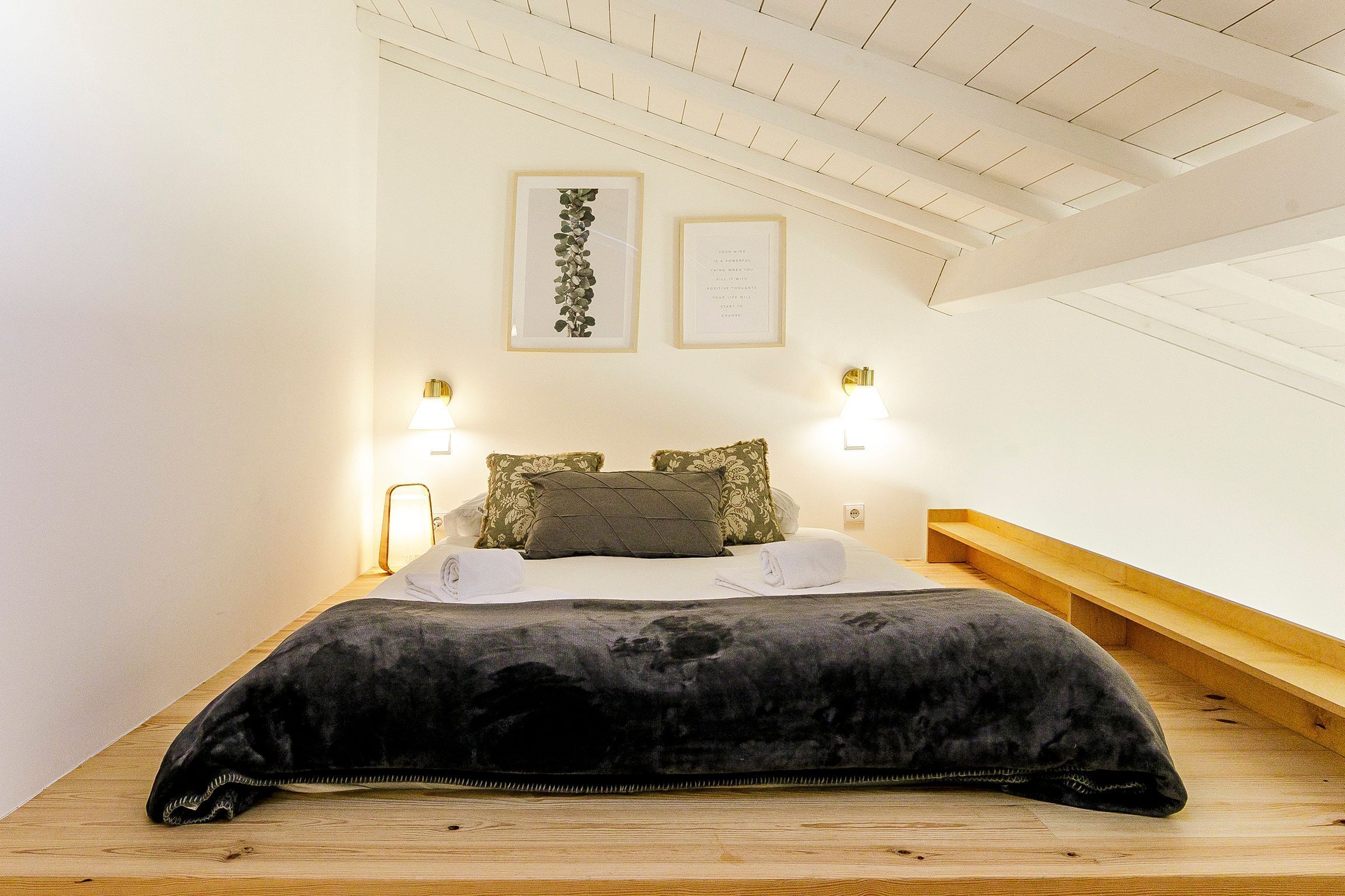 Une chambre à coucher avec un lit, deux lampes fixées au mur, une estampe botanique encadrée et un banc en bois.