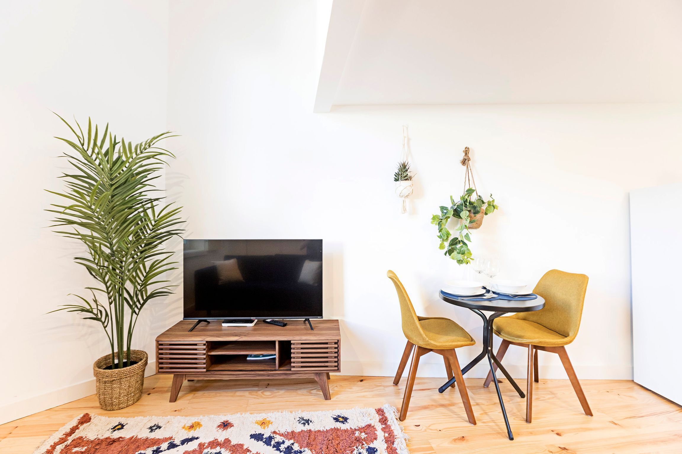 A living room with a flat-screen TV on a wooden stand, a potted plant, two yellow chairs, and a small dining table with a white tablecloth.