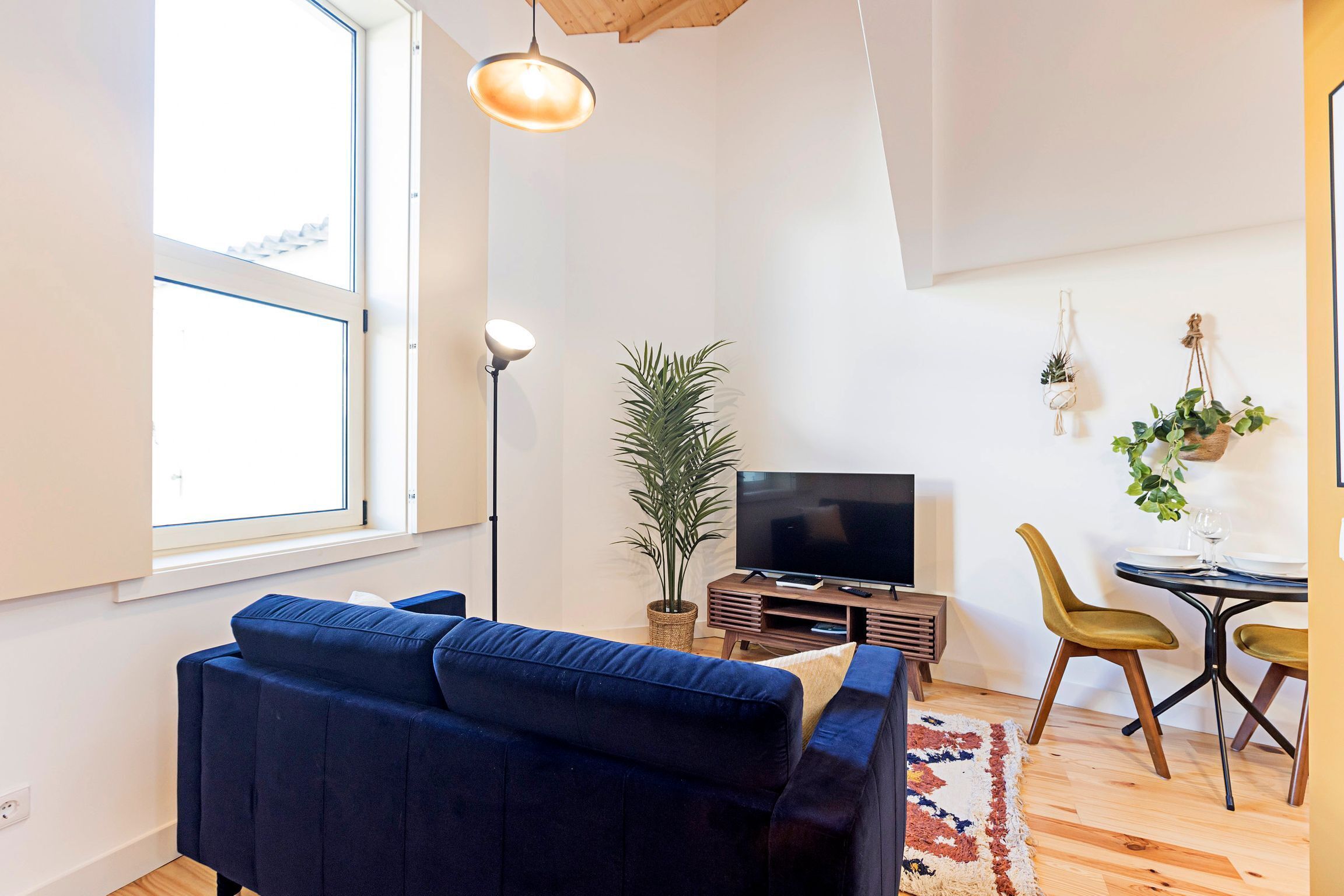 A modern living room with a blue sofa, a flat-screen TV, a floor lamp, and a dining area with yellow chairs and a small table.
