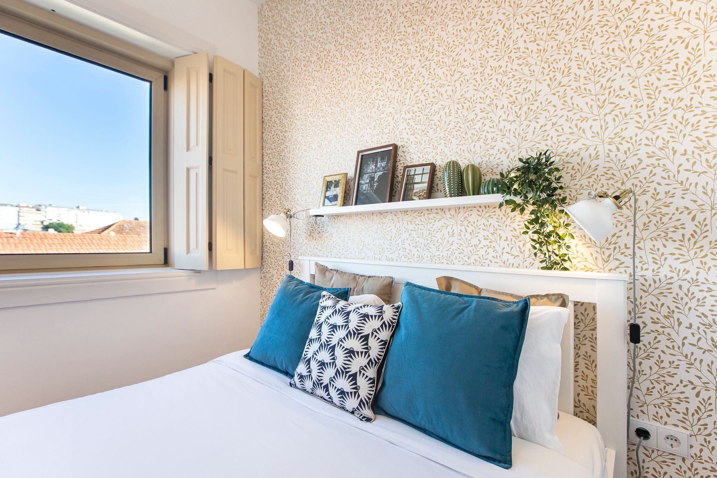 A bedroom with a bed, pillows, a nightstand, a shelf with decorative items, a window, and a wall with a leafy pattern.