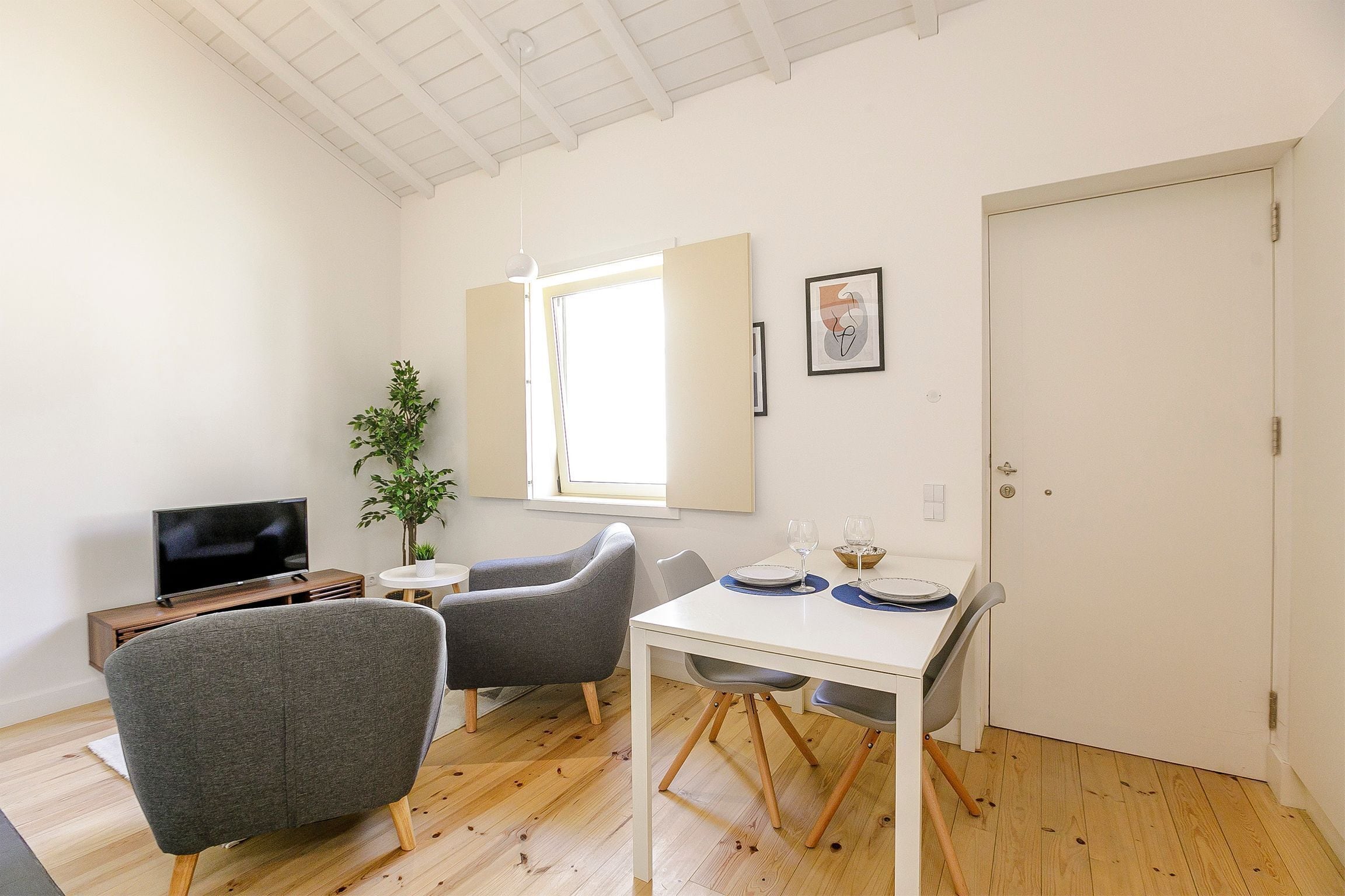 Un salon moderne doté d’un téléviseur à écran plat, de fauteuils gris, d’une table à manger blanche avec des sets de table bleus et d’une grande fenêtre laissant entrer la lumière naturelle.