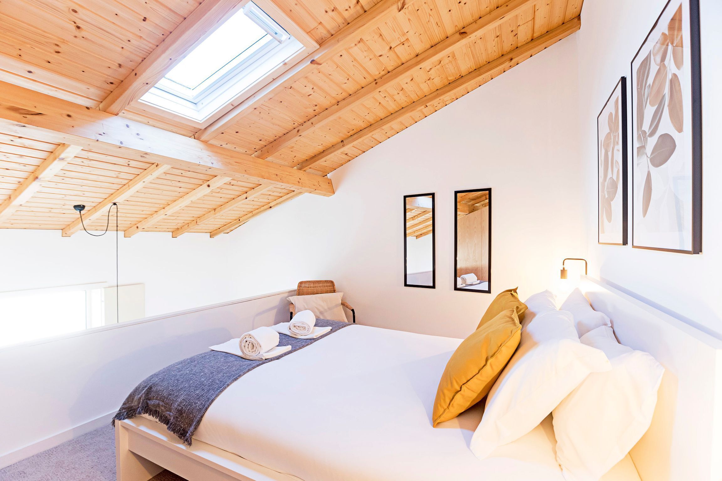 A bedroom with a sloped ceiling, skylight, bed with pillows, towels, and framed artwork on the wall.