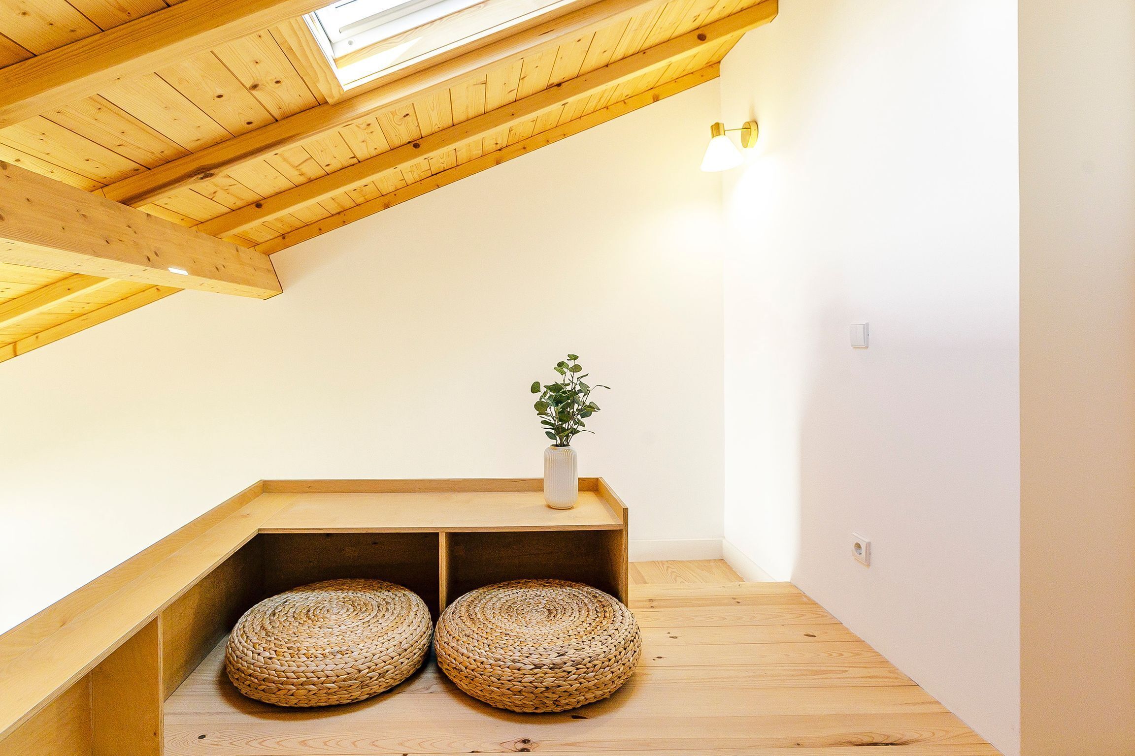 Una stanza con soffitto spiovente, travi in legno, un tavolino con un vaso e una pianta, e due pouf intrecciati sul pavimento.