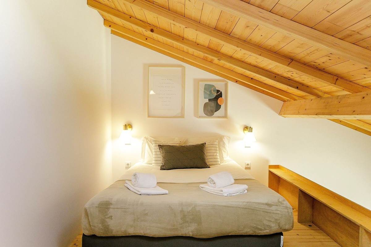 A bedroom with a wooden ceiling, a bed with two pillows, two bedside lamps, and two rolled towels on the bed.
