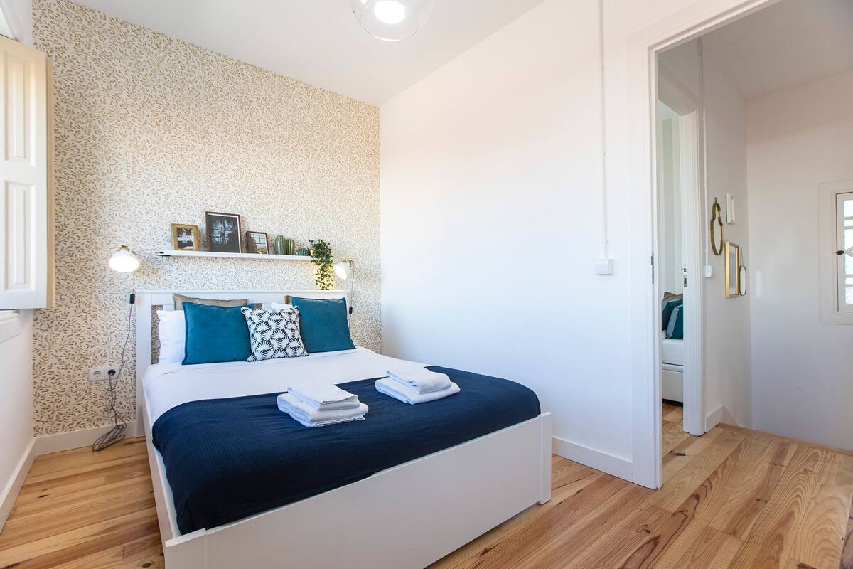 A bedroom with a bed, a shelf with decorative items, a bedside lamp, and a wooden floor.