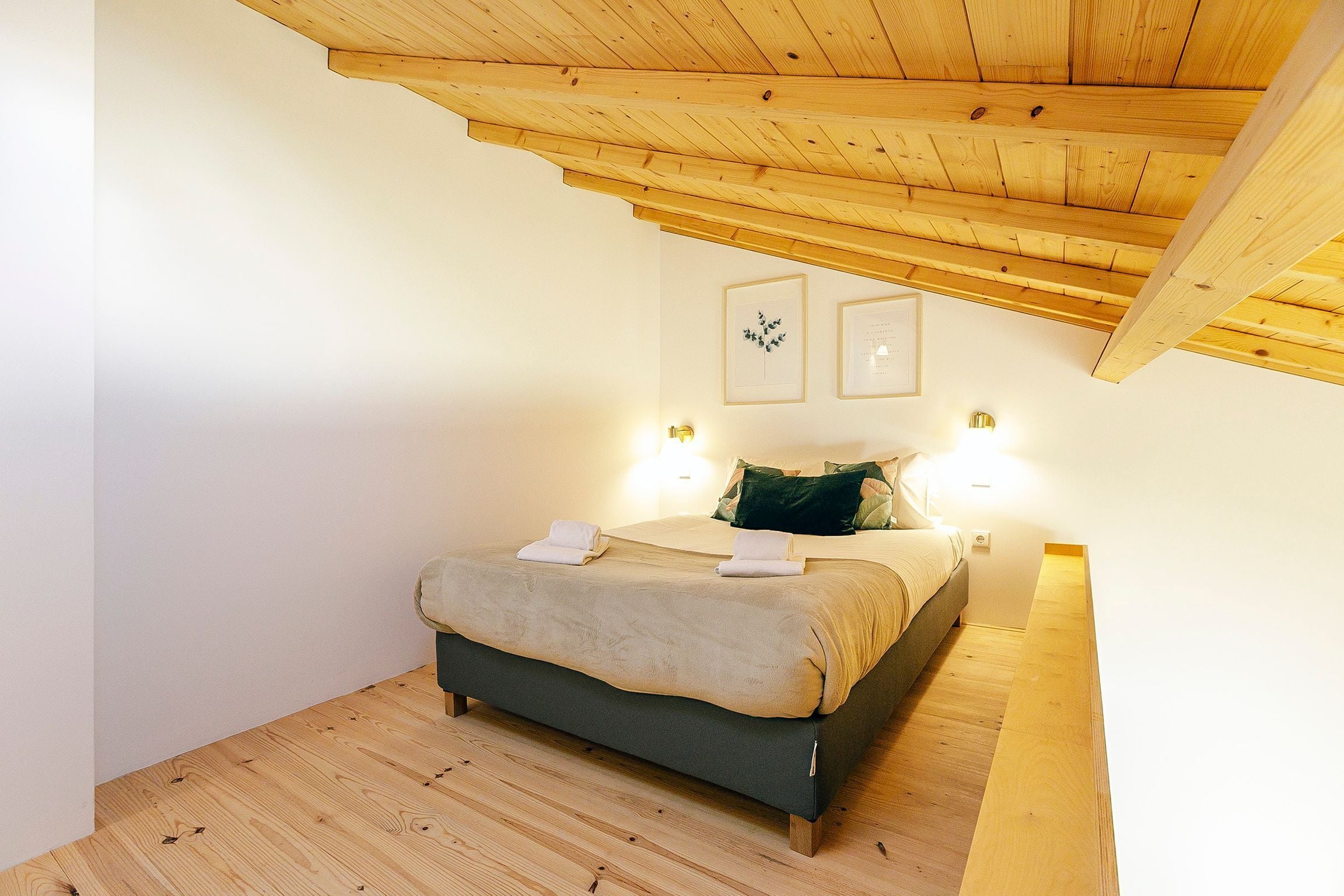 Une chambre avec un plafond en bois, un lit doté d’une tête de lit sombre, deux lampes fixées au mur et des tableaux encadrés accrochés au mur.