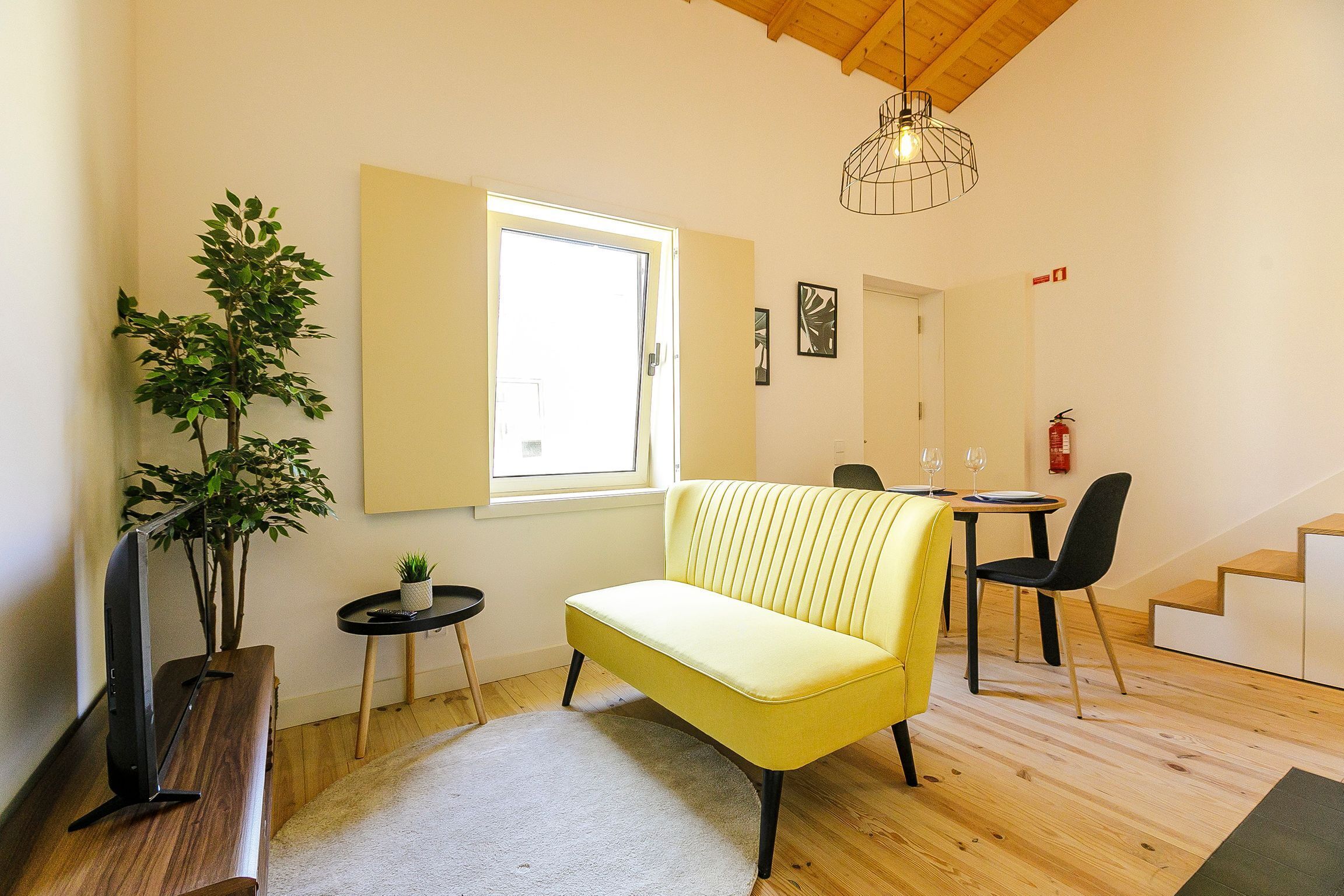 A modern living room with a yellow sofa, a wooden TV stand, a small side table, a potted plant, and a large window.
