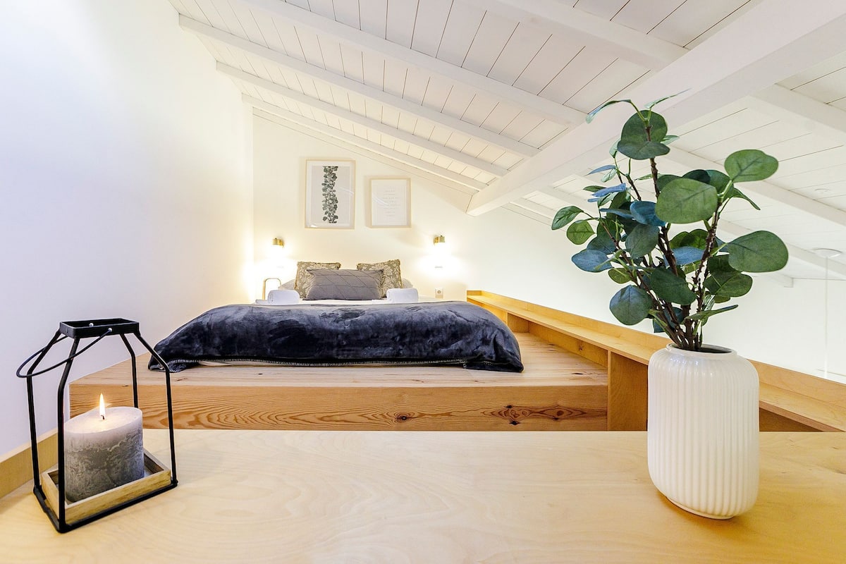 A bedroom with a bed, a bedside table, a nightstand, a large potted plant, and a wall-mounted shelf.